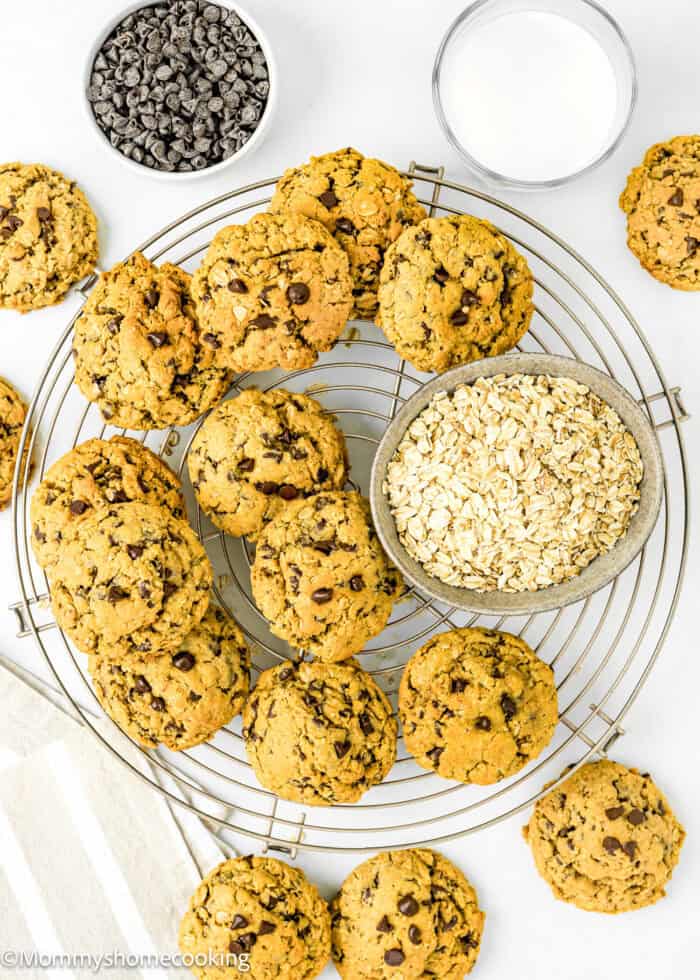 baked Egg-free Oatmeal Chocolate Chip Cookies over a cooling rack with a bowl of chocolate chips, oats, a kitchen towel and a glass of milk on the sides.