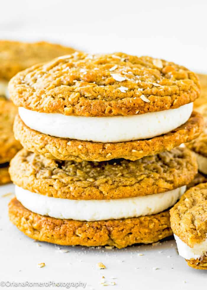 Close-up of stacked eggless oatmeal cream pies filled with vanilla ice cream, with additional cookies in the background.