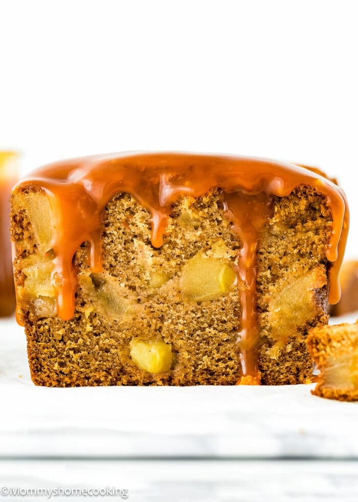 A close-up of a glazed loaf cake with visible chunks of fruit inside. A slice of this eggless, easy apple quick bread is cut and placed next to the cake.