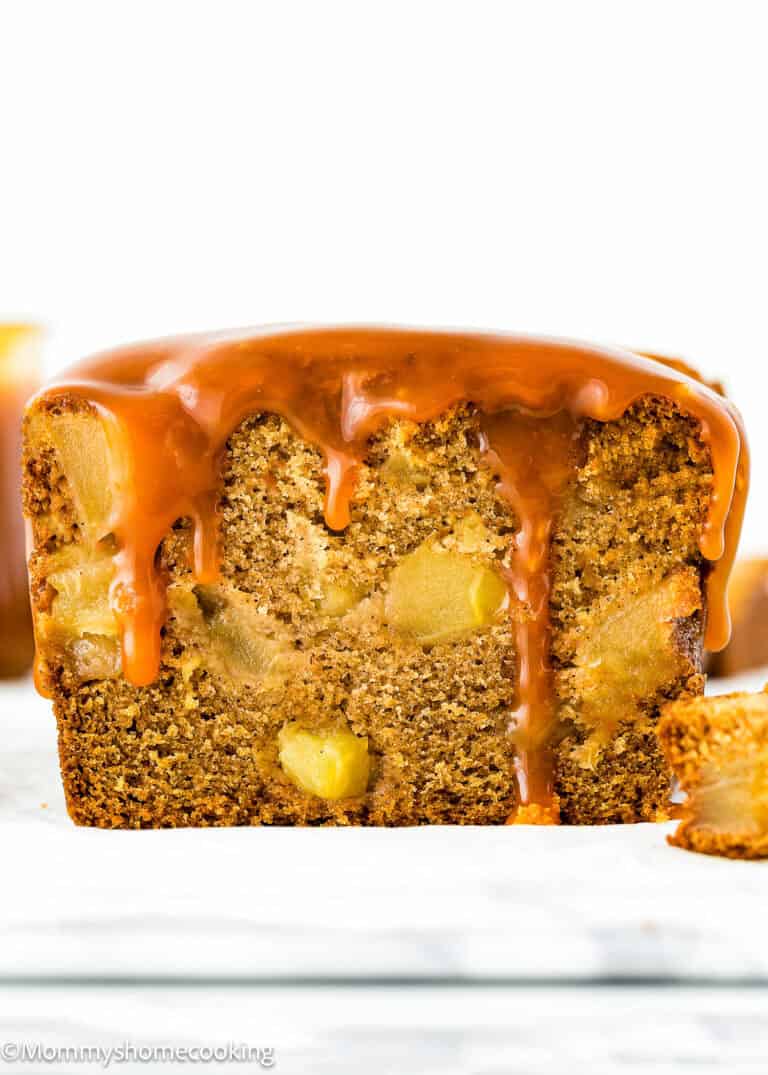 A close-up of a glazed loaf cake with visible chunks of fruit inside. A slice of this eggless, easy apple quick bread is cut and placed next to the cake.