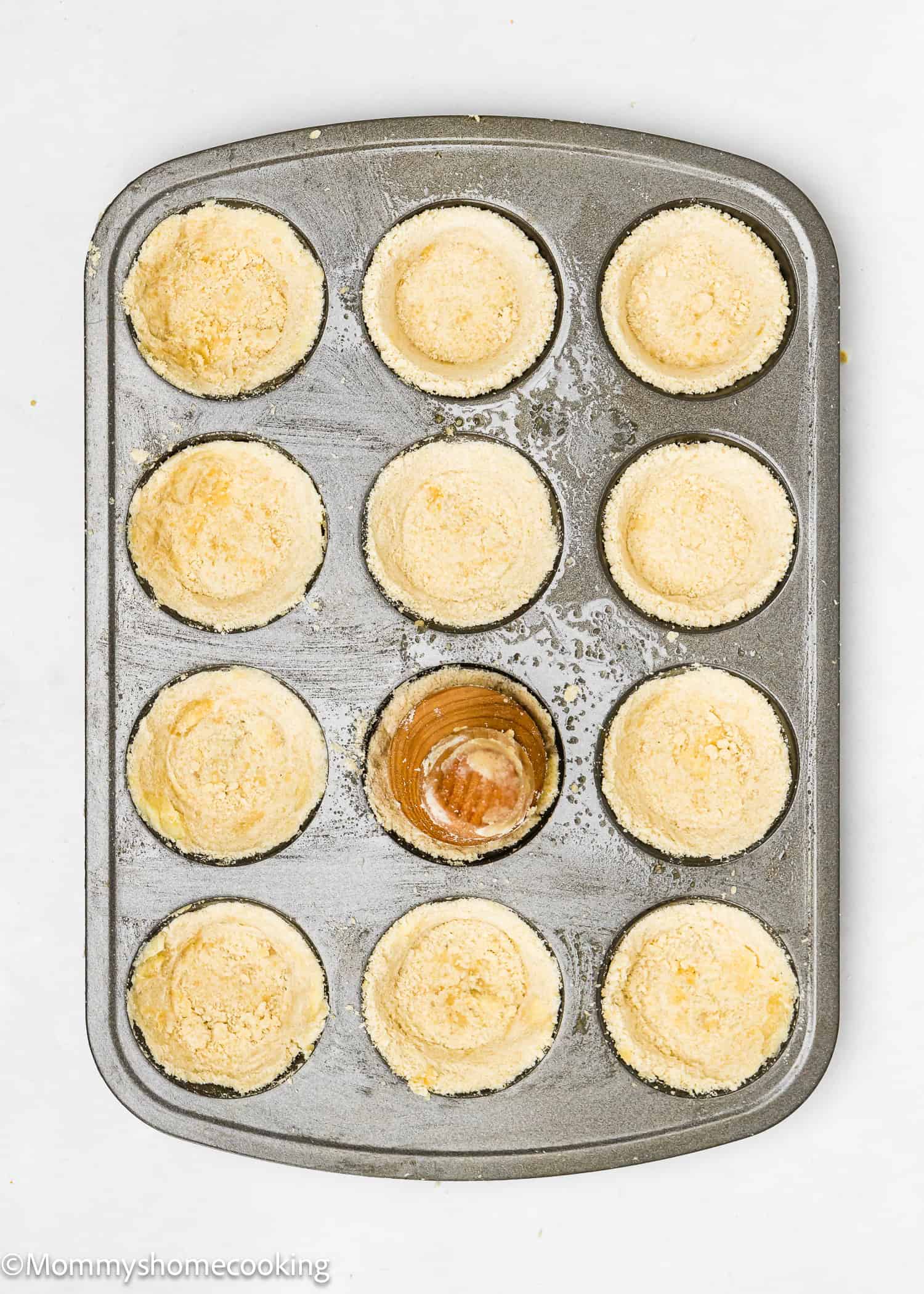 A muffin tin containing 12 compartments is filled with partially baked crusts. One compartment showcases a golden brown tart, hinting at the delicious promise of eggless mini pumpkin pies.