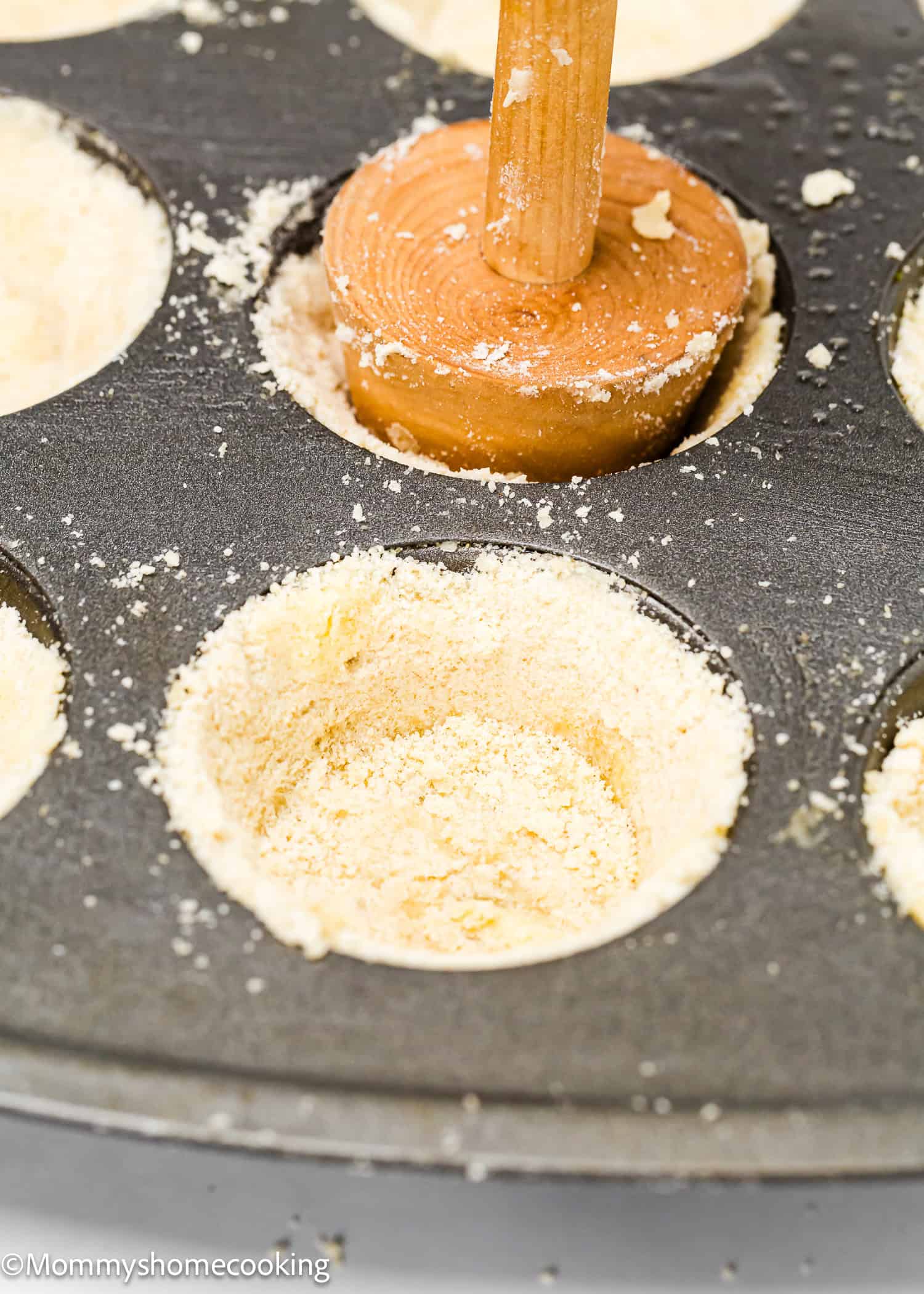 Using a wooden tamper, carefully press the shortbread dough into each mini muffin pan cavity, creating the perfect base for eggless mini pumpkin pies.