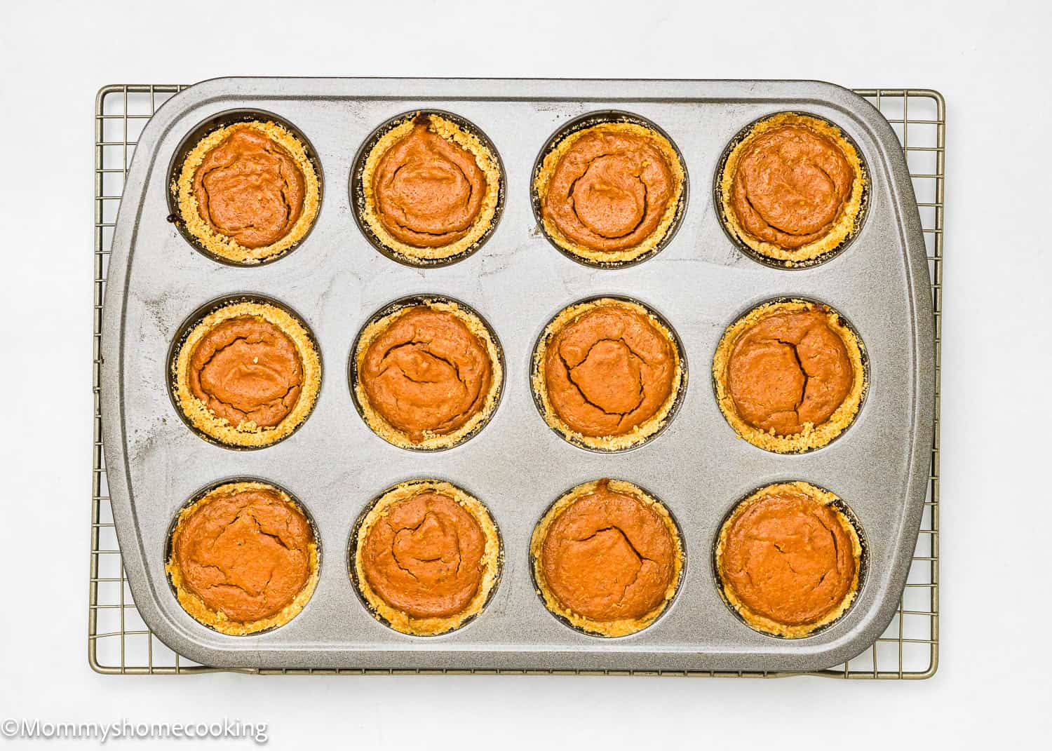 A muffin tin with twelve freshly baked eggless mini pumpkin pies cools on a wire rack.