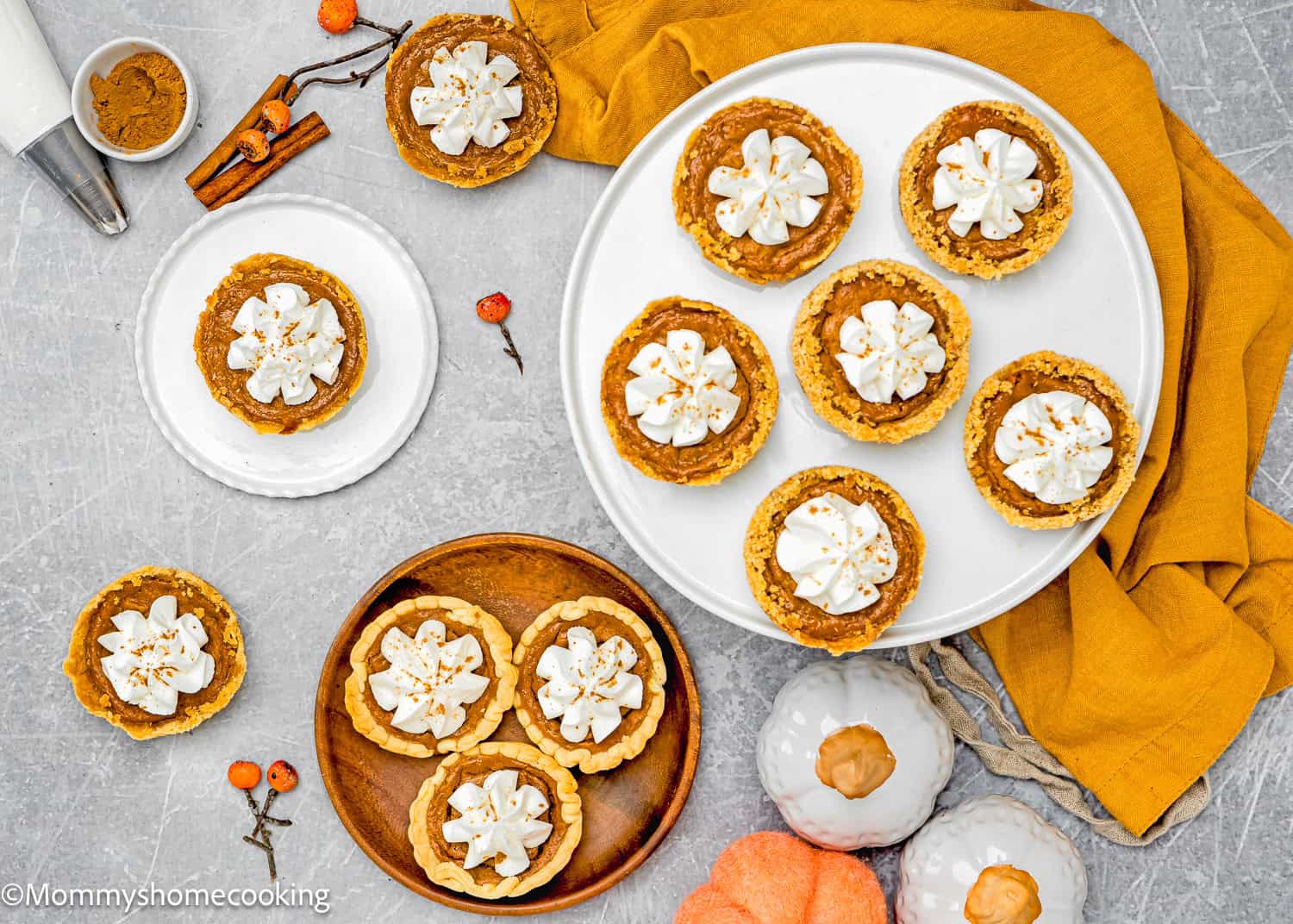 Top view of eggless mini pumpkin pies topped with whipped cream on a table, surrounded by fabric, small decorations, and pumpkins.