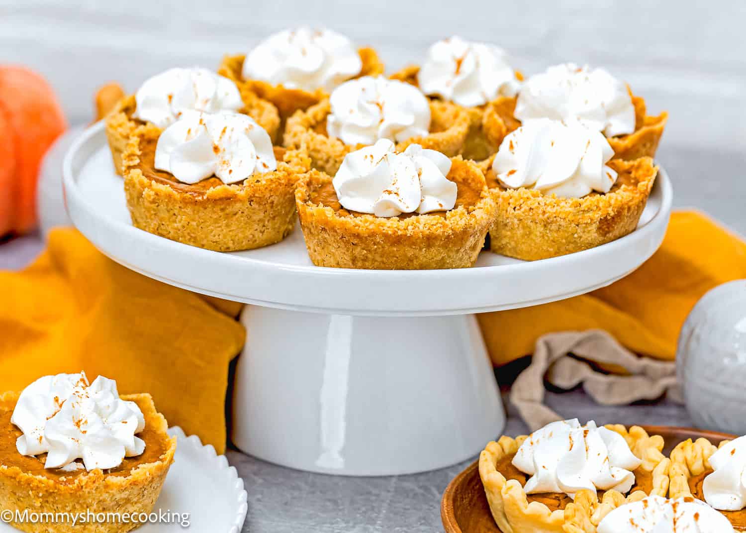 Eggless mini pumpkin pies topped with whipped cream are artfully arranged on a white cake stand, surrounded by a yellow cloth and a perfectly placed pumpkin.