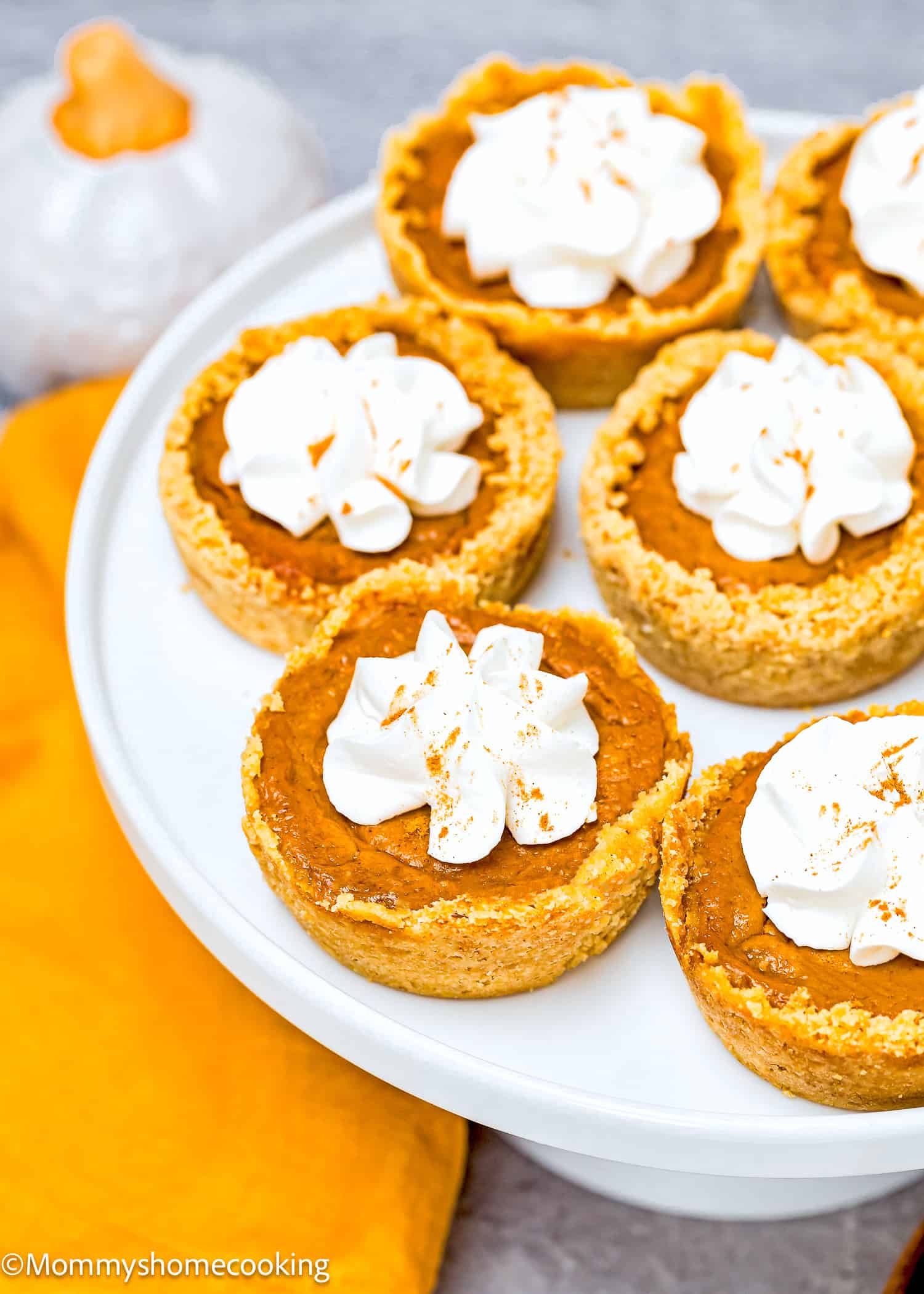 Eggless mini pumpkin pies topped with whipped cream sit elegantly on a white cake stand, with an orange cloth and a small decorative pumpkin in the background.