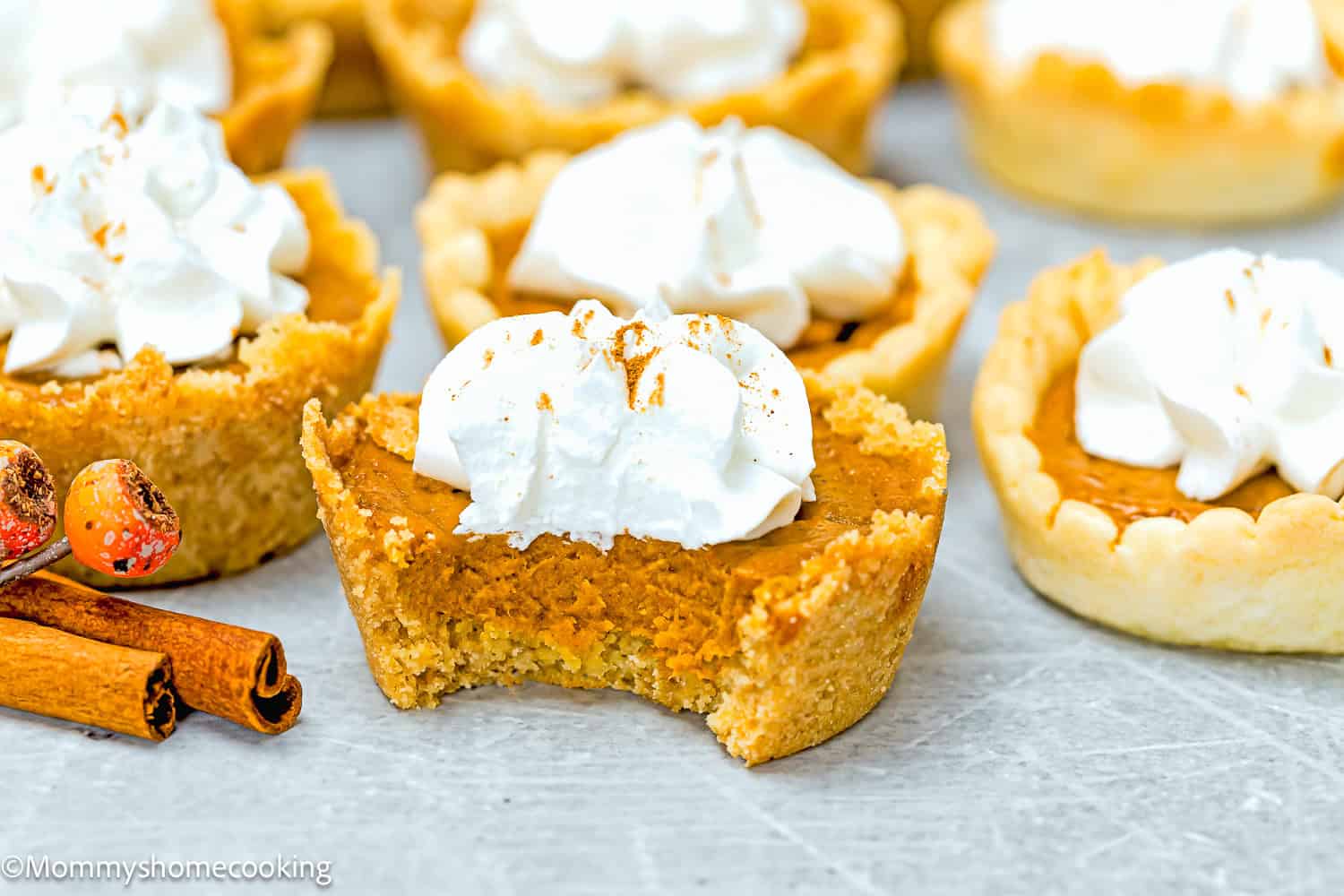 Eggless mini pumpkin pies with whipped cream sit on a gray surface, one showcasing a tempting bite. Nearby, cinnamon sticks and fall decorations evoke the cozy warmth of the season.