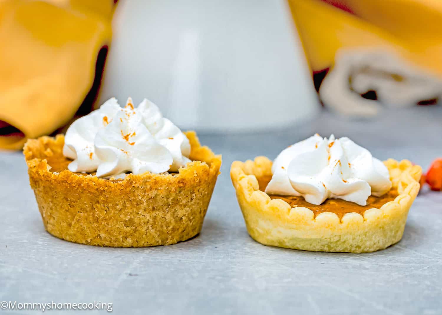 Two eggless mini pumpkin pies topped with whipped cream sit on a gray surface, with a blurred background of yellow fabric.