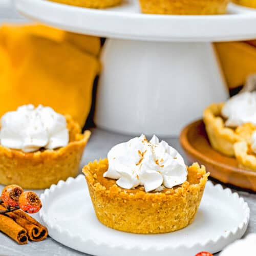 Eggless mini pumpkin pies topped with whipped cream rest on a white plate, with more mouthwatering delights elegantly displayed in the background on a cake stand.