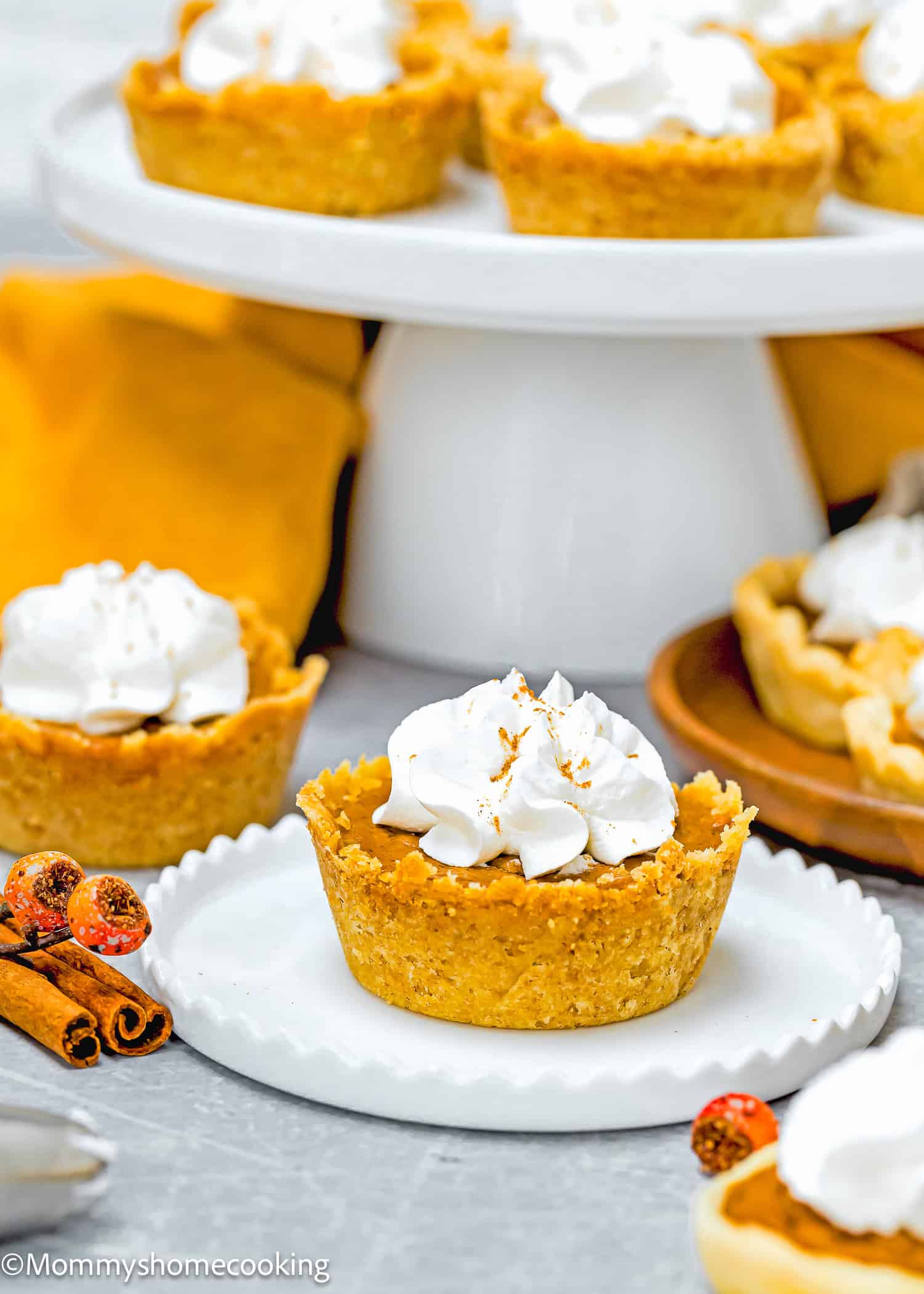 Eggless mini pumpkin pies topped with whipped cream rest on a white plate, with more mouthwatering delights elegantly displayed in the background on a cake stand.