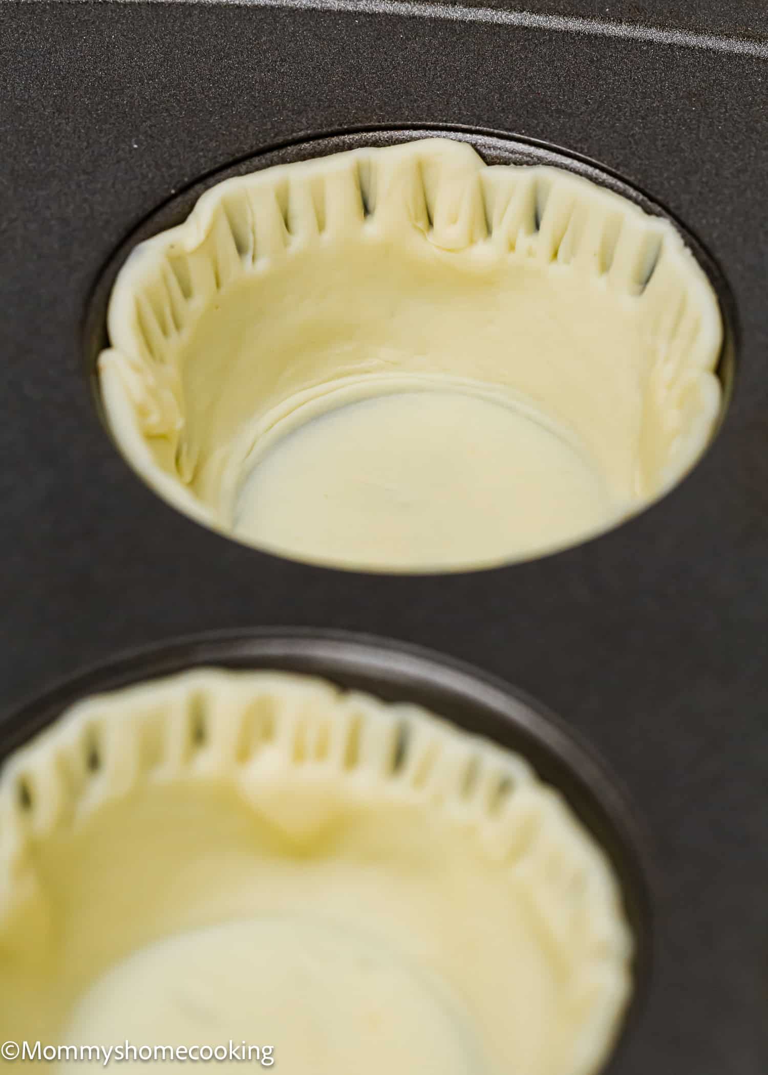 Close-up of a muffin tin with pastry dough pressed into two of the cups, ready for baking delightful eggless mini pumpkin pies.