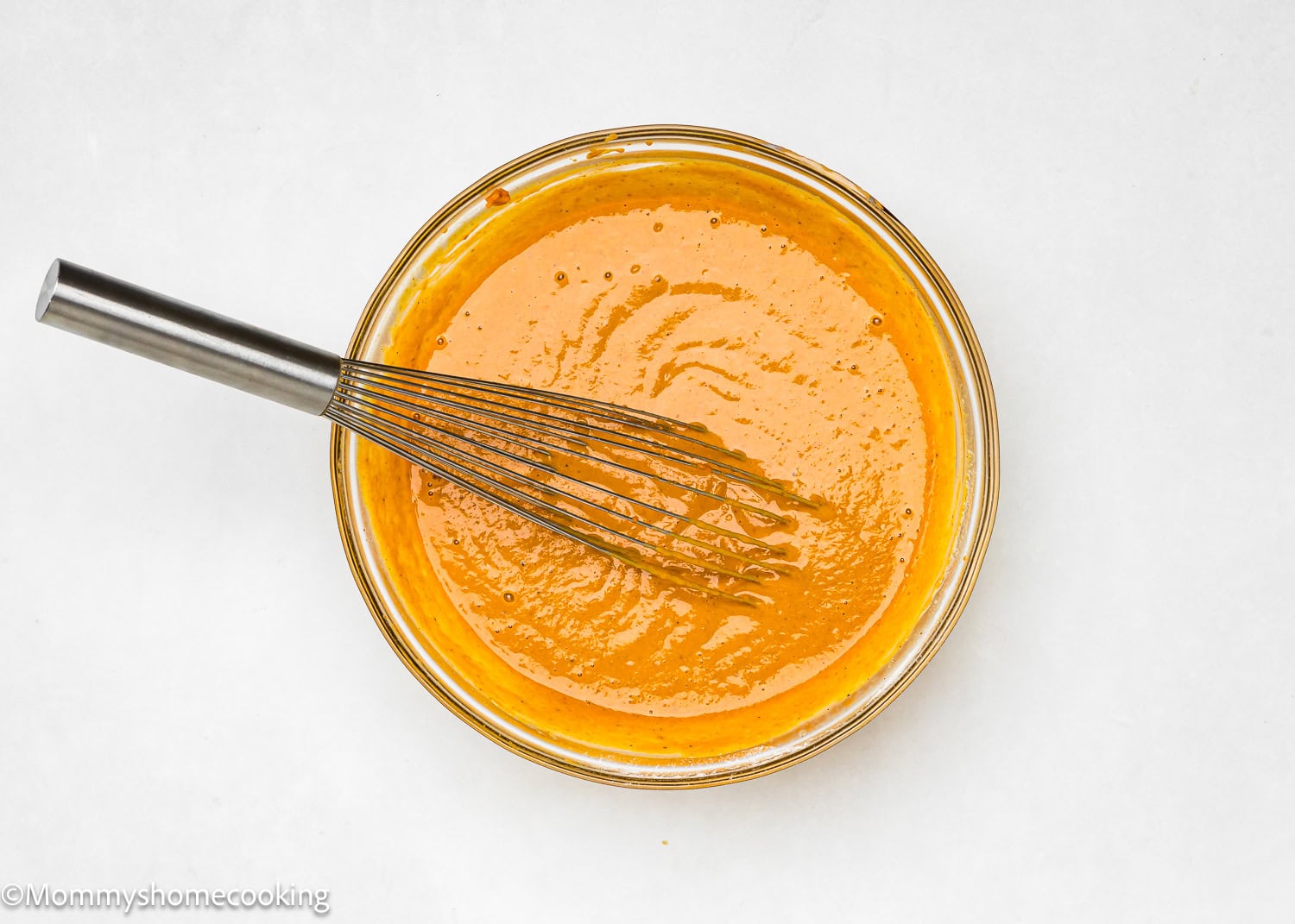 An immaculate glass bowl cradles vibrant orange batter, ready to transform into delightful eggless mini pumpkin pies. A sturdy metal whisk accompanies the scene against a pristine white backdrop, inviting you to imagine the delicious creations that await.