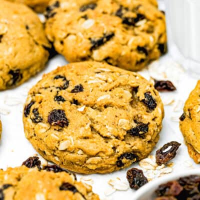 Close-up of one bowl Eggless Oatmeal Raisin Cookies on a white surface, surrounded by scattered oats and raisins, showcasing just how easy they are to make.