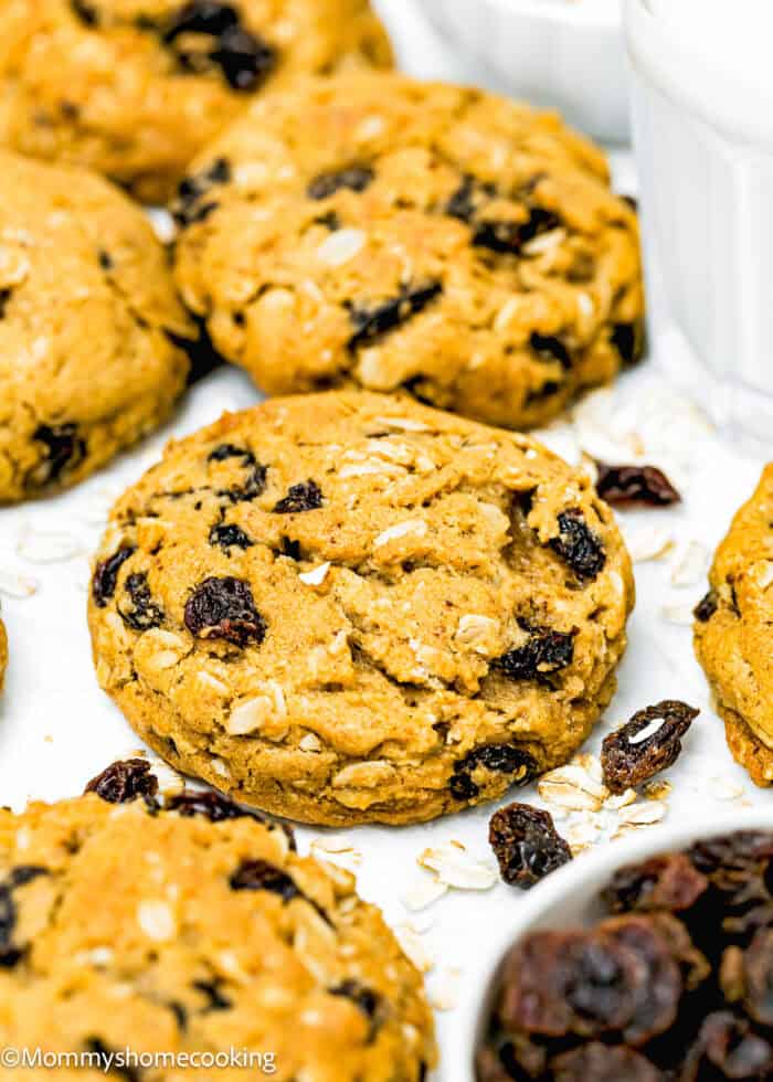 Close-up of one bowl Eggless Oatmeal Raisin Cookies on a white surface, surrounded by scattered oats and raisins, showcasing just how easy they are to make.