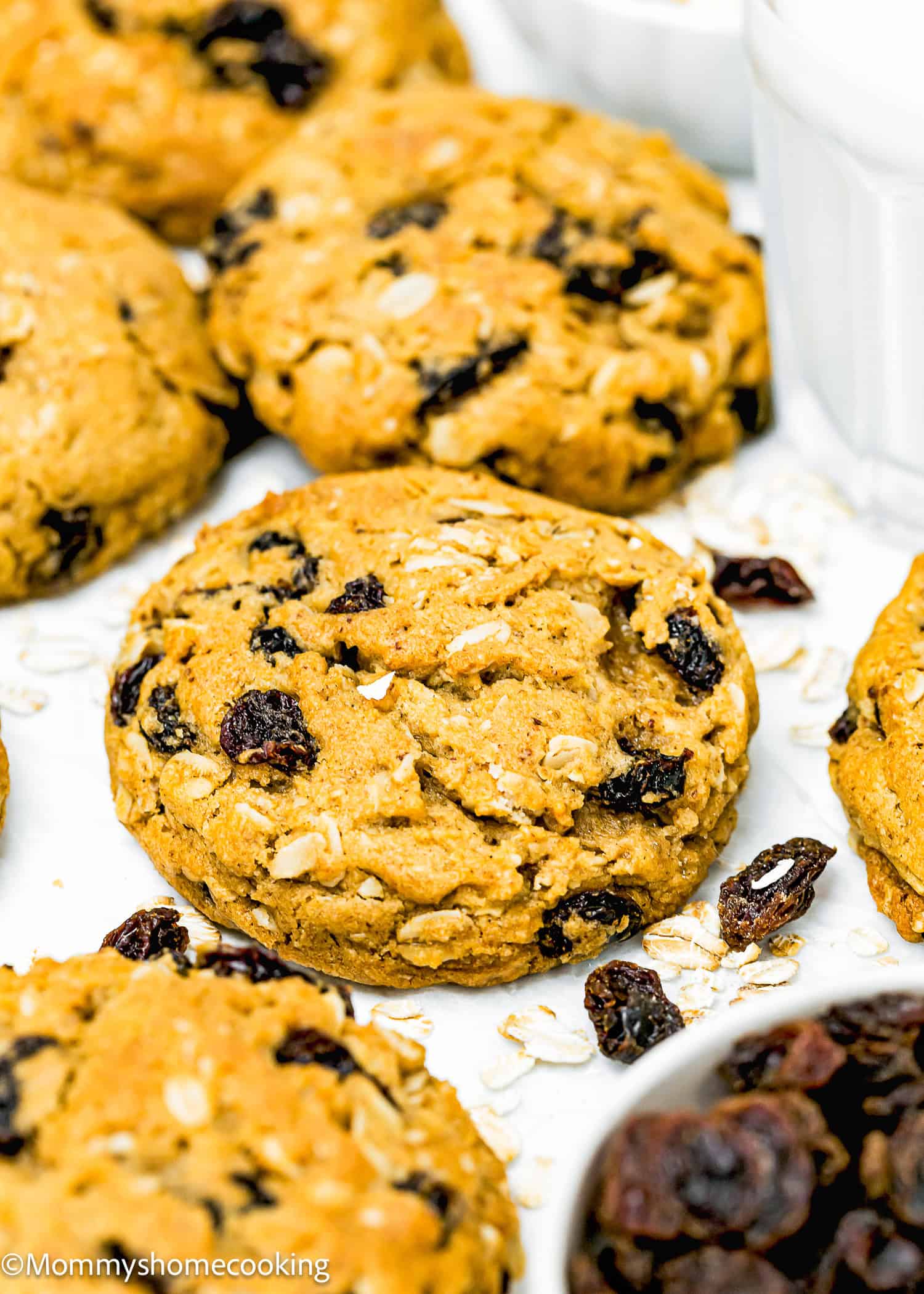 Close-up of one bowl Egg-free Oatmeal Raisin Cookies on a white surface, surrounded by scattered oats and raisins, showcasing just how easy they are to make.