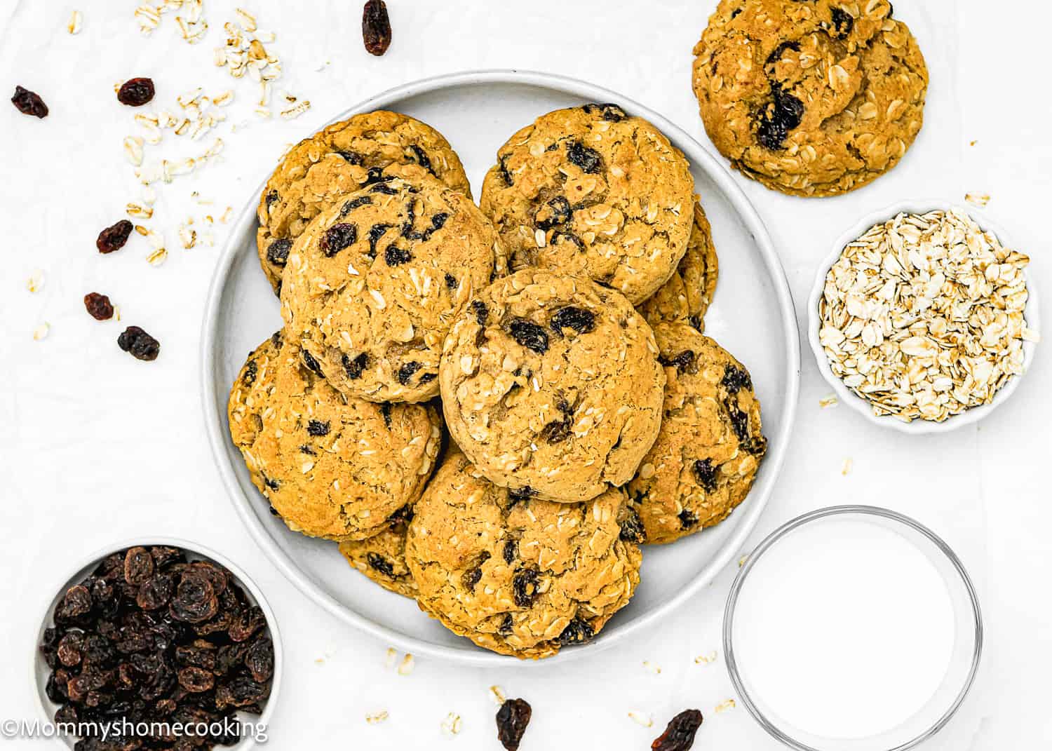 A plate of Eggless Oatmeal Raisin Cookies, surrounded by raisins, oats, and a glass of milk on a white background, exemplifies how these one-bowl treats are easy to make and deliciously indulgent.