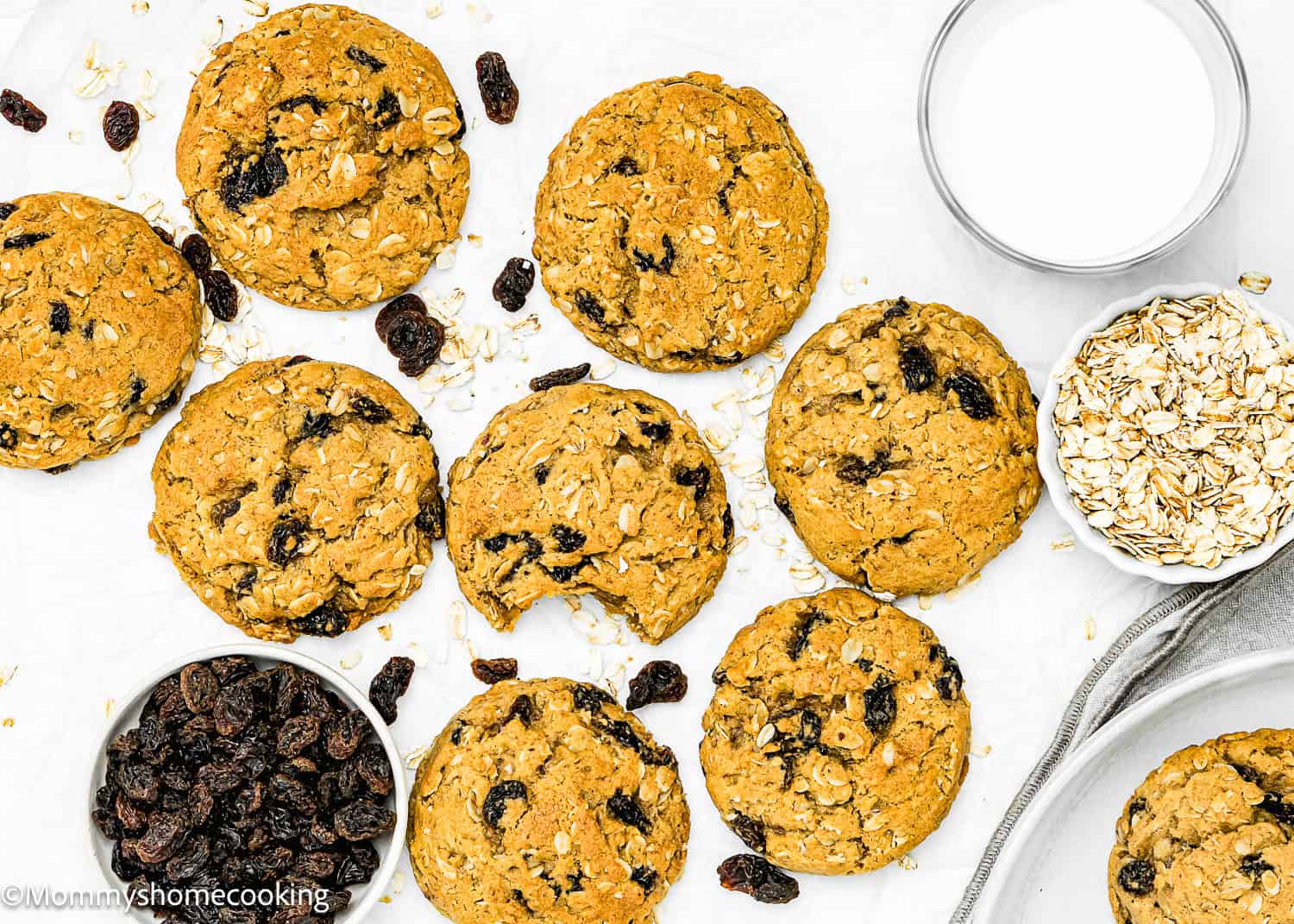 A batch of Oatmeal Raisin Cookies sits on a white surface, surrounded by a glass of milk and bowls filled with oats and raisins. One cookie, easy to make, has a bite taken out.