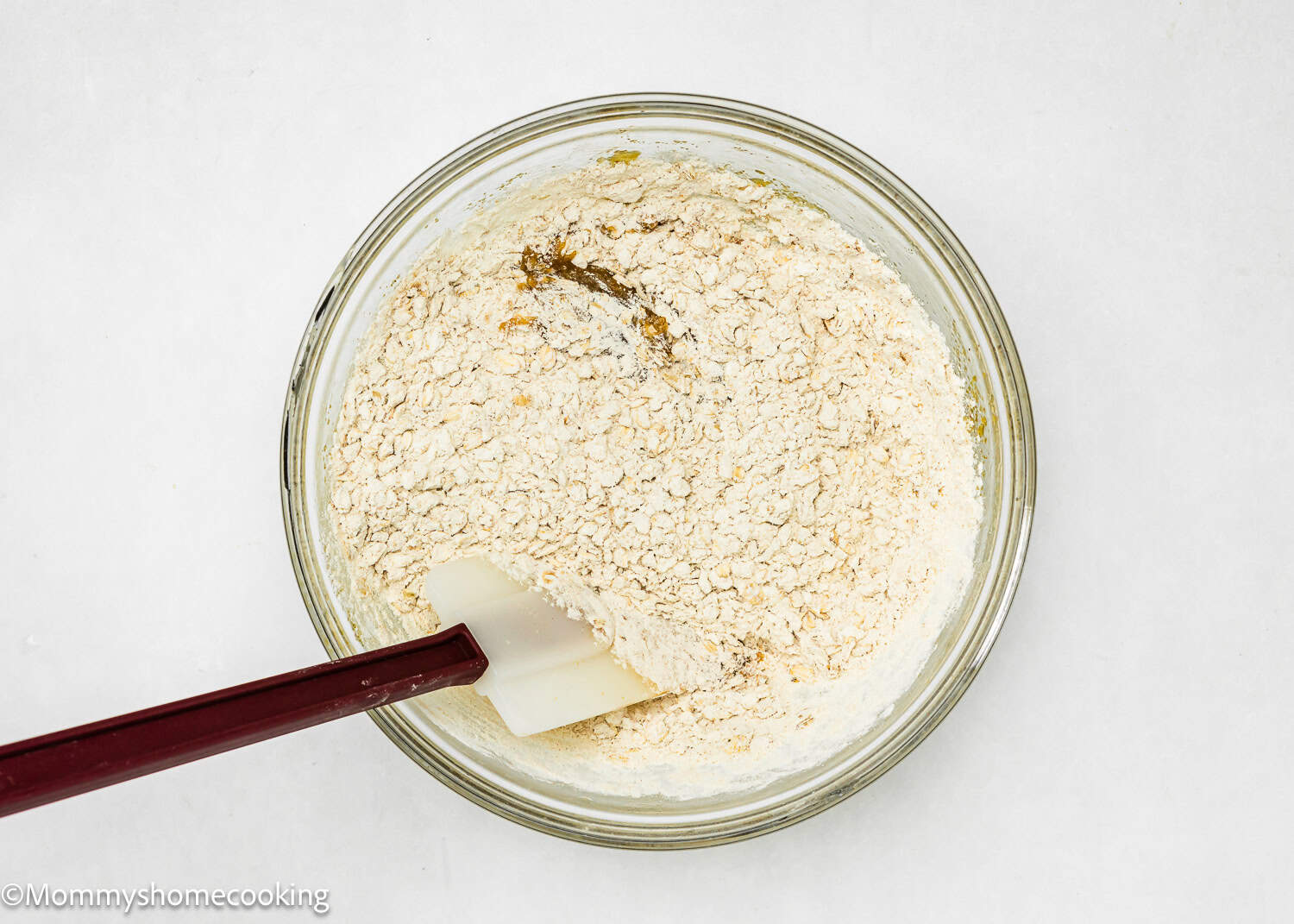 A glass bowl with dry and wet ingredients and a rubber spatula on a white surface, perfect for creating easy-to-make, eggless oatmeal raisin cookies in just one bowl.