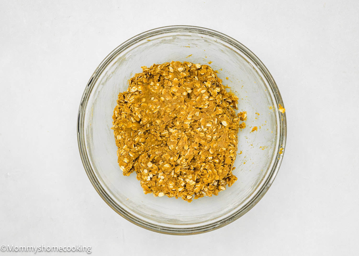 A glass bowl sits on a white surface, holding the easy-to-make, eggless oatmeal raisin cookie mixture.