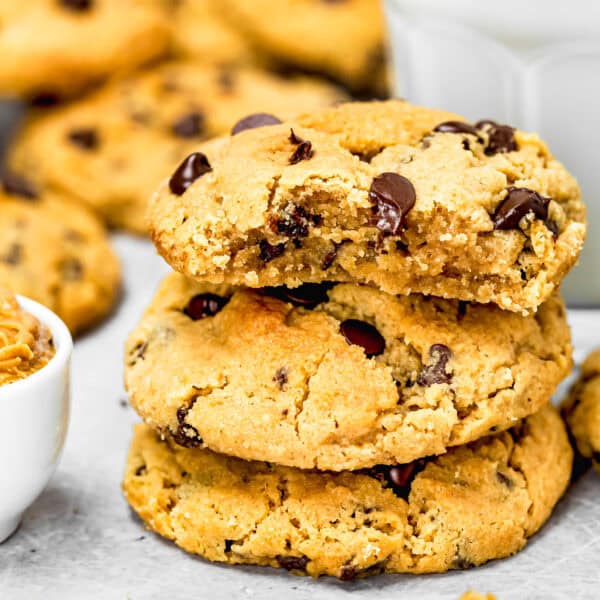 A stack of three eggless peanut butter chocolate chip cookies with a bite taken out of the top one. More cookies and a glass of milk are visible in the background.