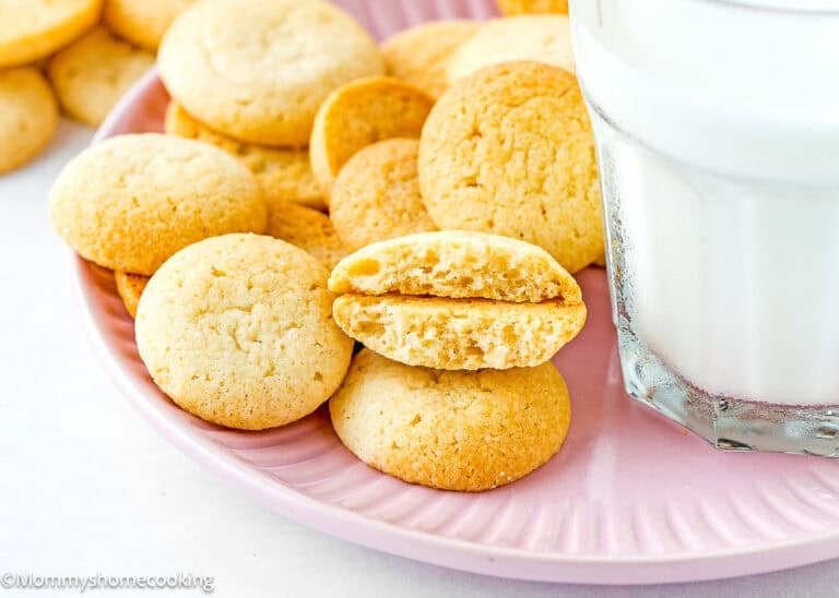A pink plate with several round eggless vanilla wafers, one broken in half, sits next to a glass of milk.