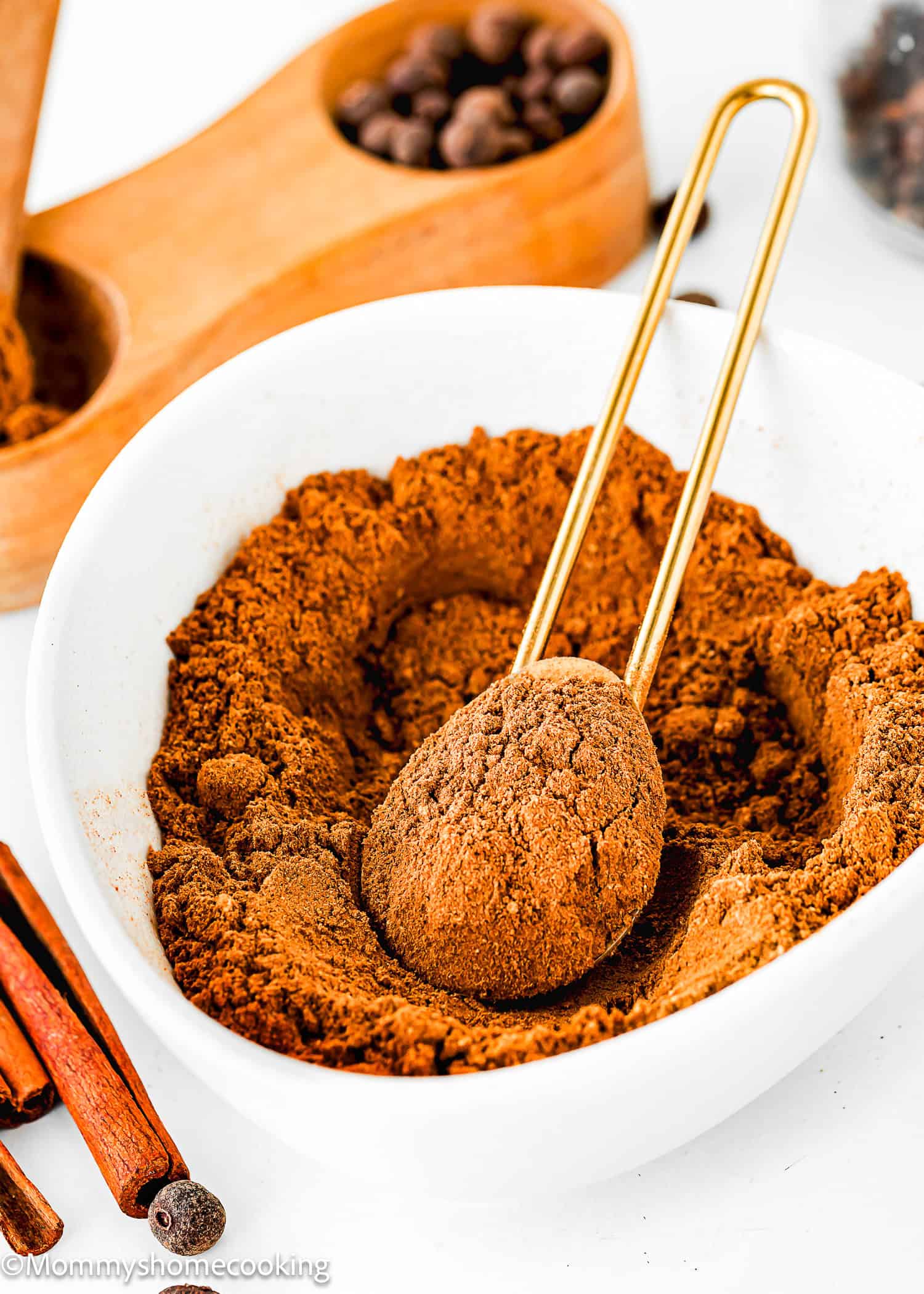 A bowl of homemade pumpkin pie spice blend with a gold spoon, cinnamon sticks, and nutmeg visible in the background.