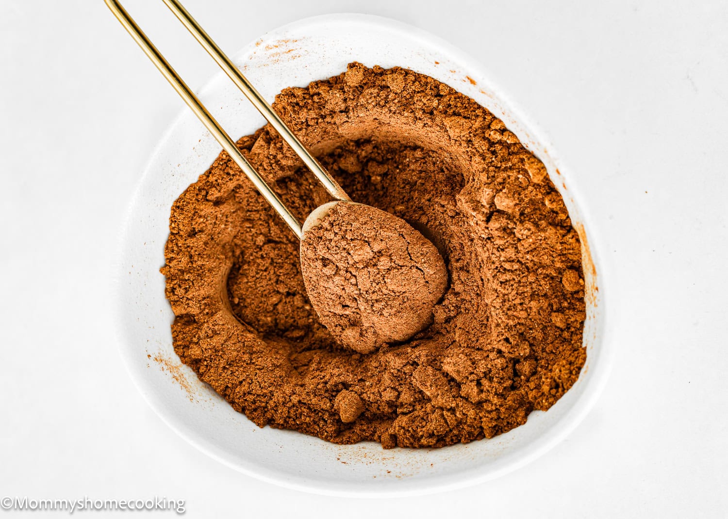 A heart-shaped bowl, filled with rich cocoa powder, sits invitingly beside two gold measuring spoons, ready for crafting a homemade pumpkin pie spice blend.