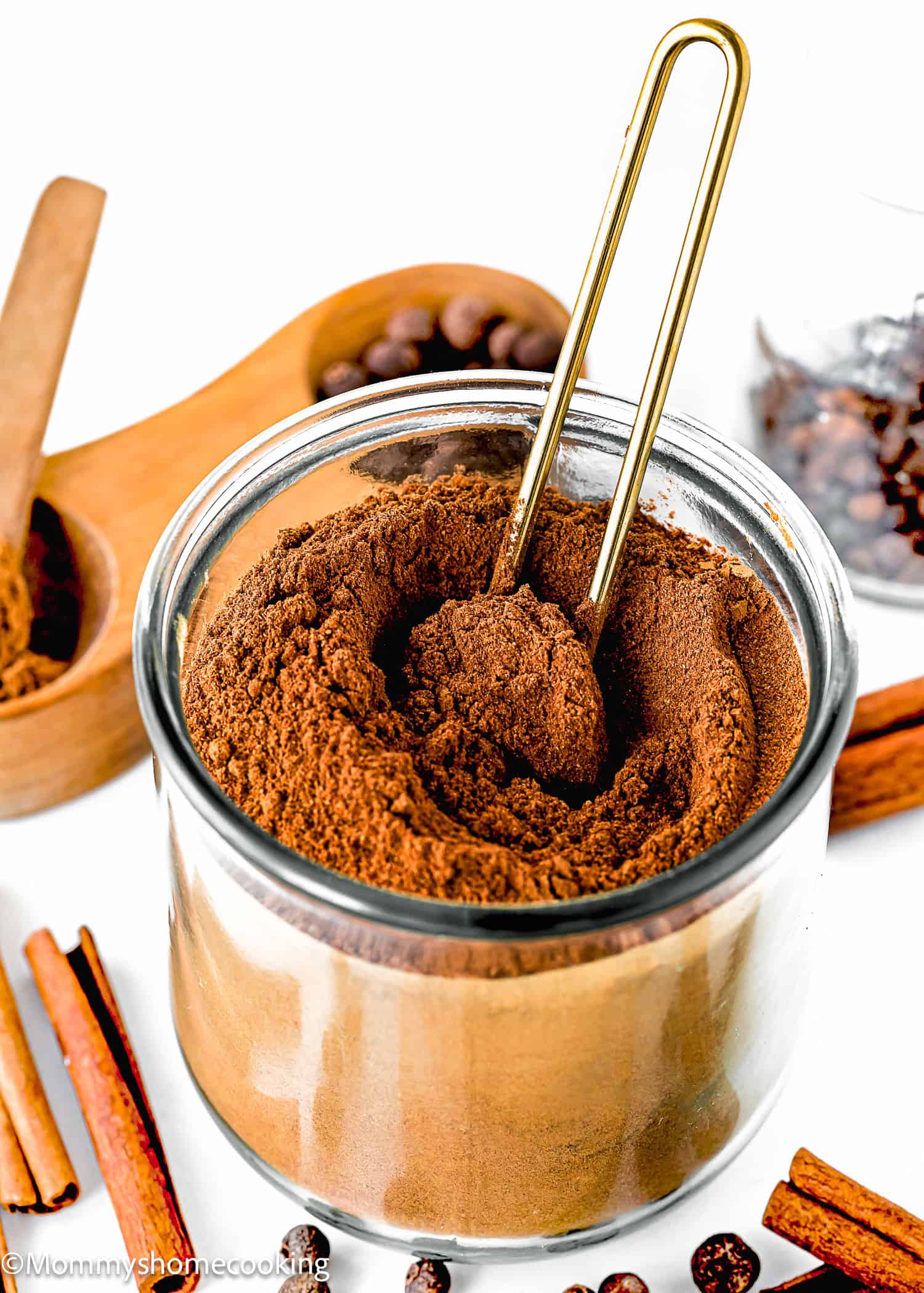 A jar of cocoa powder with a gold scoop sits amidst cinnamon sticks, coffee beans, and a wooden container of homemade pumpkin pie spice blend.