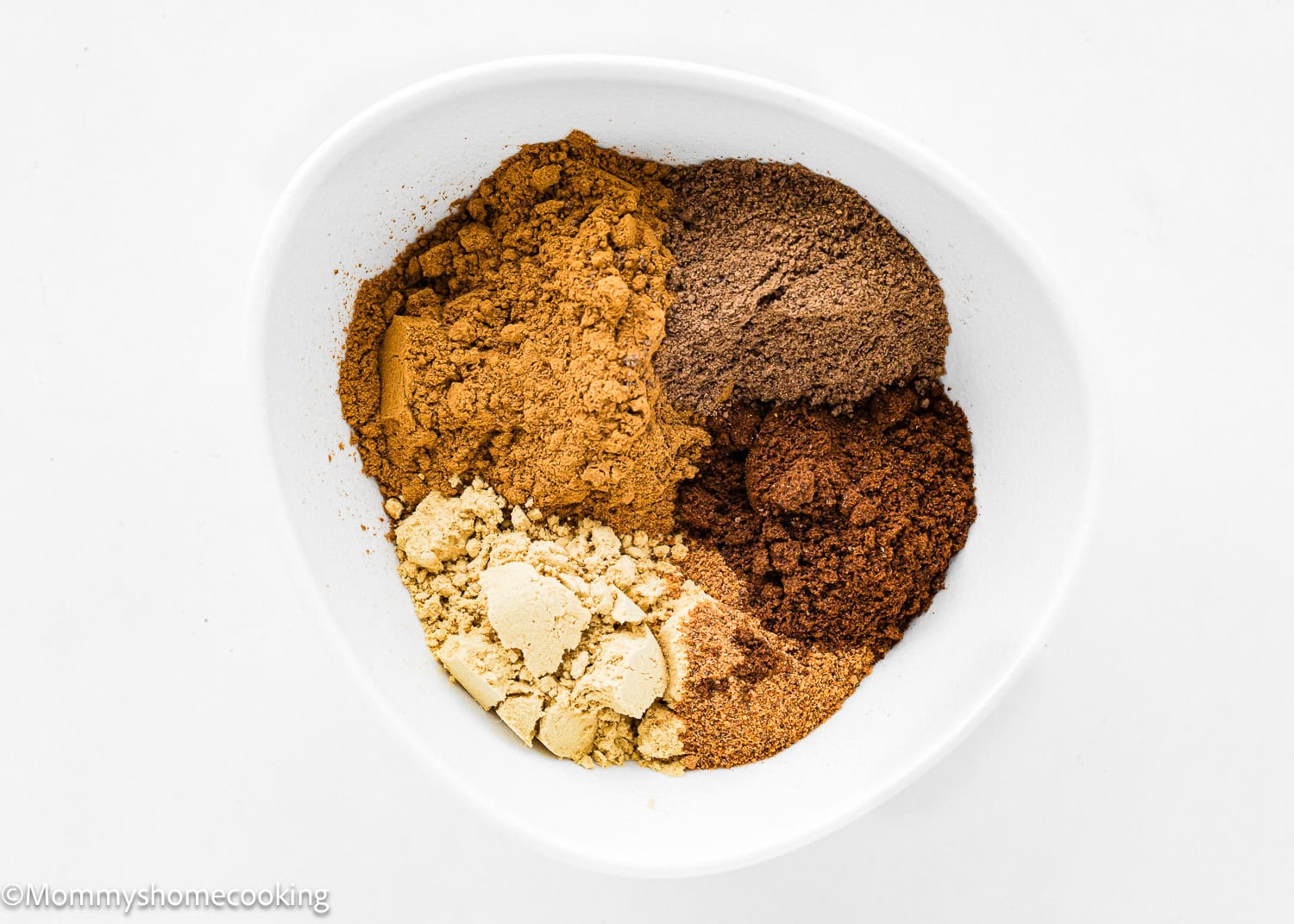 A bowl showcases a homemade pumpkin pie spice blend with five distinct brown and beige spices neatly arranged in separate sections on a pristine white background.