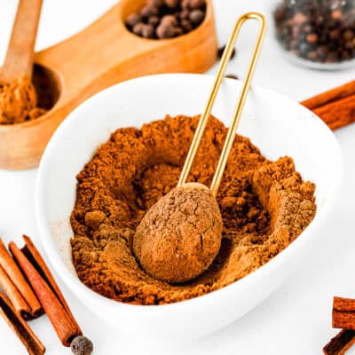 A white bowl filled with homemade pumpkin pie spice blend and a measuring spoon. Surrounding the bowl are cinnamon sticks and whole spices, creating an inviting aroma.