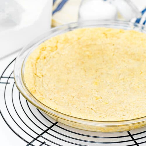 A no-fuss, gluten-free pie crust sits in a clear pie dish on a cooling rack, surrounded by baking materials. This easy press-in pie crust requires no chill, making your baking experience even simpler.