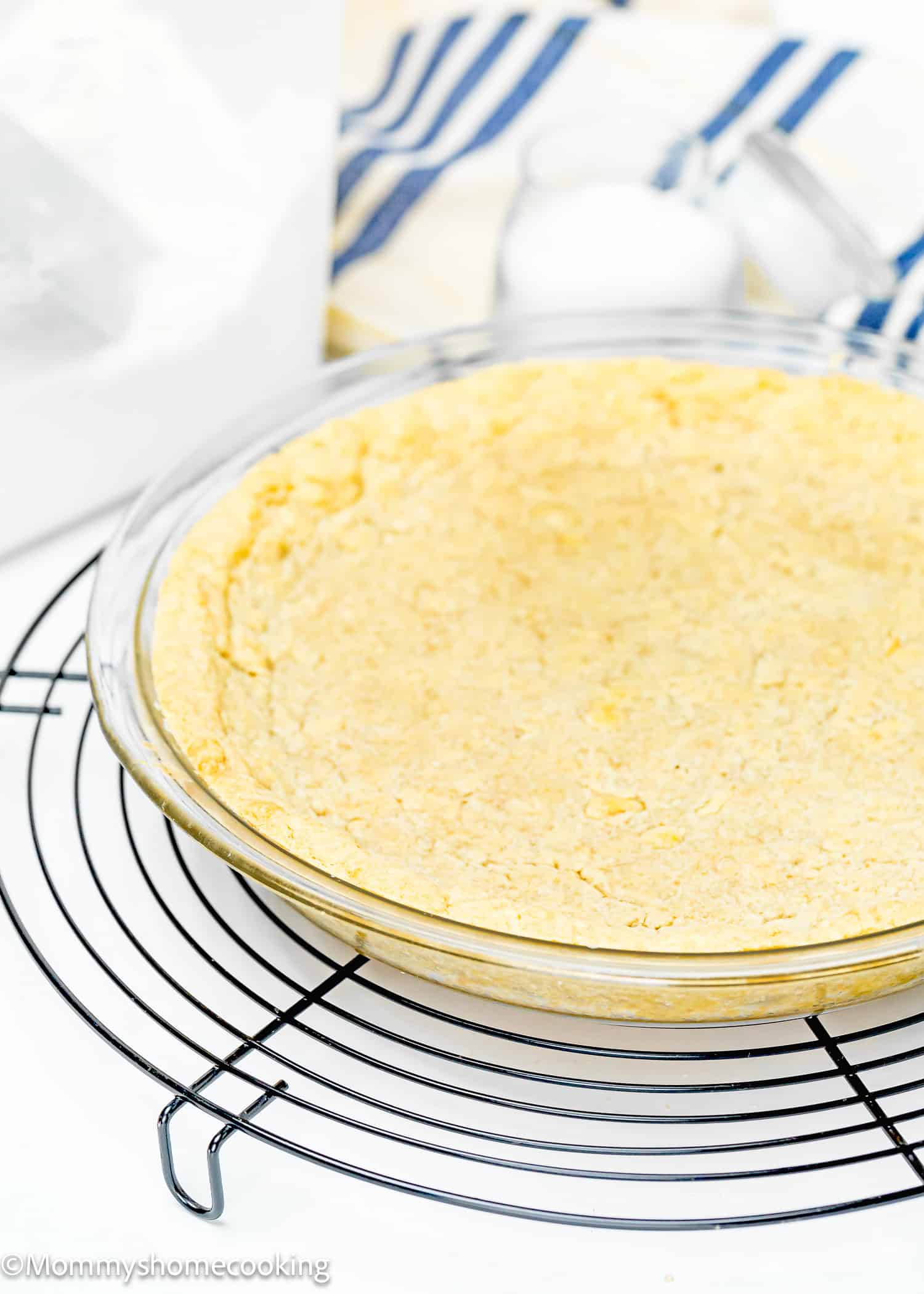 A no-fuss, gluten-free pie crust sits in a clear pie dish on a cooling rack, surrounded by baking materials. This easy press-in pie crust requires no chill, making your baking experience even simpler.