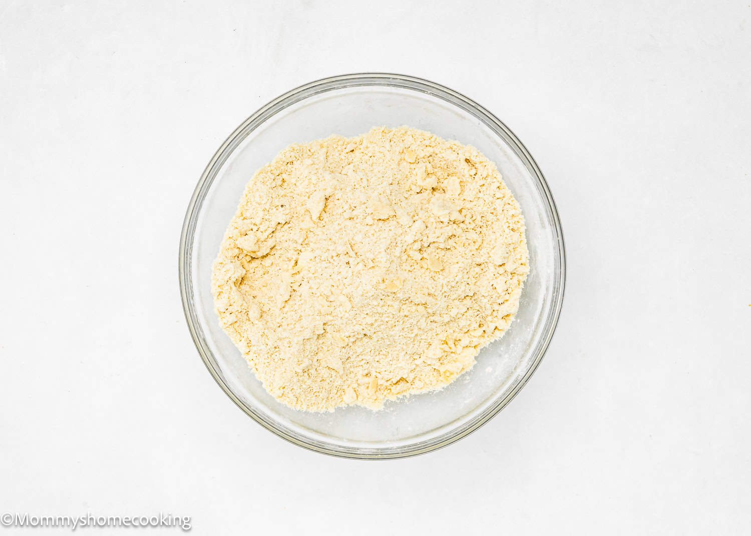 A clear glass bowl holds a heap of beige, powdery flour, perfect for an easy, no-fuss press-in pie crust against a plain white background.