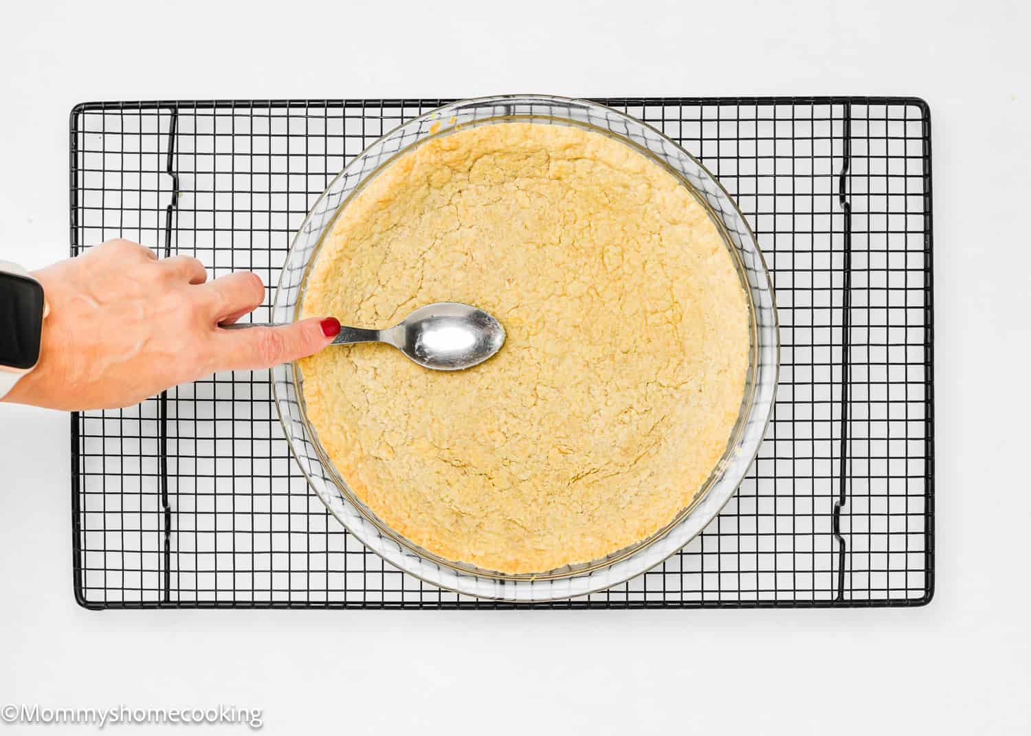 A hand with red nail polish uses a spoon to press down on the No-Fuss, Easy Press In Pie Crust dough in a glass dish, placed on a cooling rack.