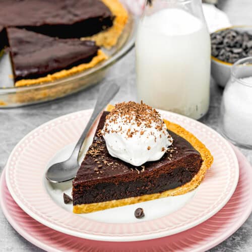 A slice of eggless chocolate pie on a pink plate with whipped cream and chocolate shavings, accompanied by a spoon, a glass of milk, and an easy brownie pie dish in the background.