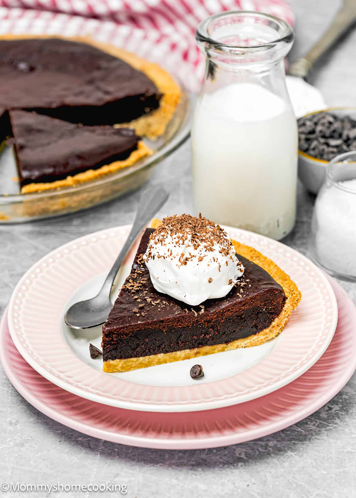 A slice of eggless chocolate pie on a pink plate with whipped cream and chocolate shavings, accompanied by a spoon, a glass of milk, and an easy brownie pie dish in the background.