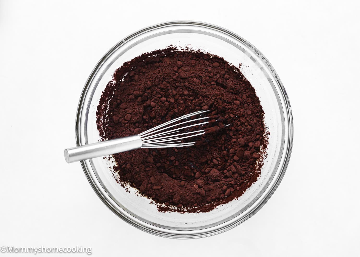 An eggless easy brownie pie mix sits in a glass bowl, with a whisk ready to blend the rich cocoa powder against a pristine white background.