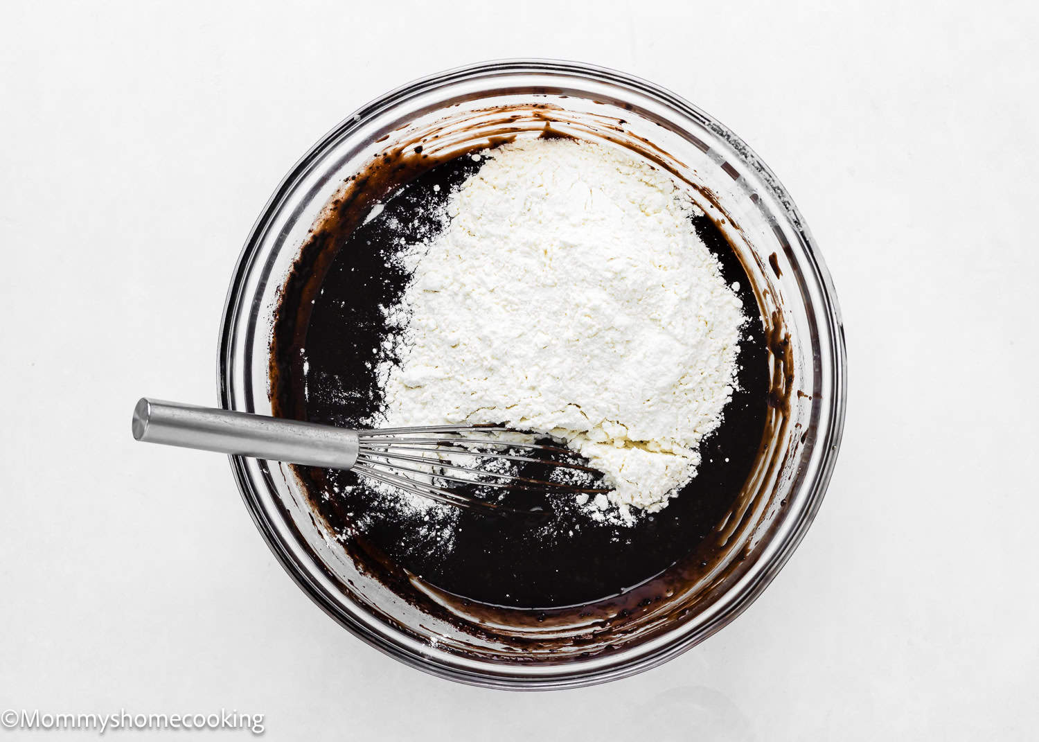 A glass bowl with a dark liquid mixture and a pile of white flour on top is being whisked, creating the smooth batter for an eggless easy brownie pie that's sure to delight.