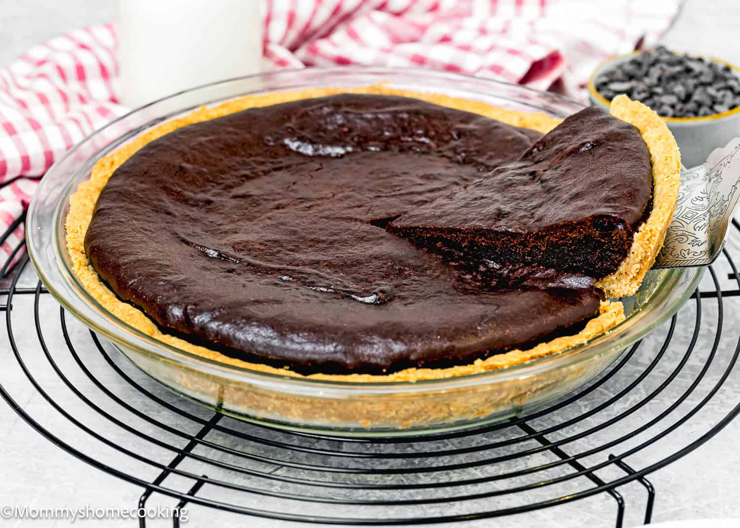 An easy brownie pie with a slice being removed, placed on a wire cooling rack. This eggless treat sits beautifully alongside a glass of milk and a small bowl of chocolate chips.