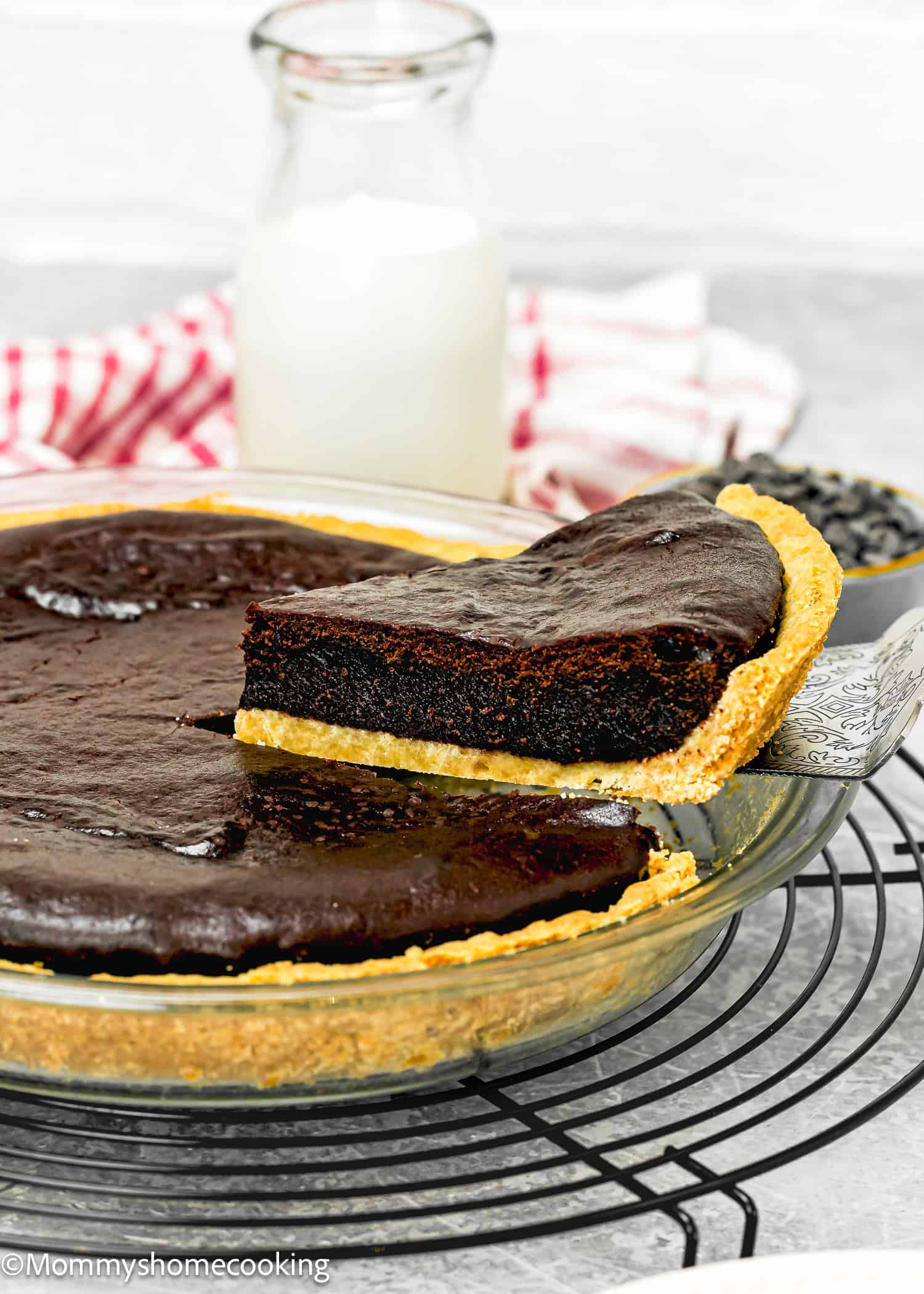 A slice of decadent dark chocolate tart, reminiscent of an eggless easy brownie pie, is being served from a glass pie dish. In the background, a bottle of milk complements this indulgent treat.