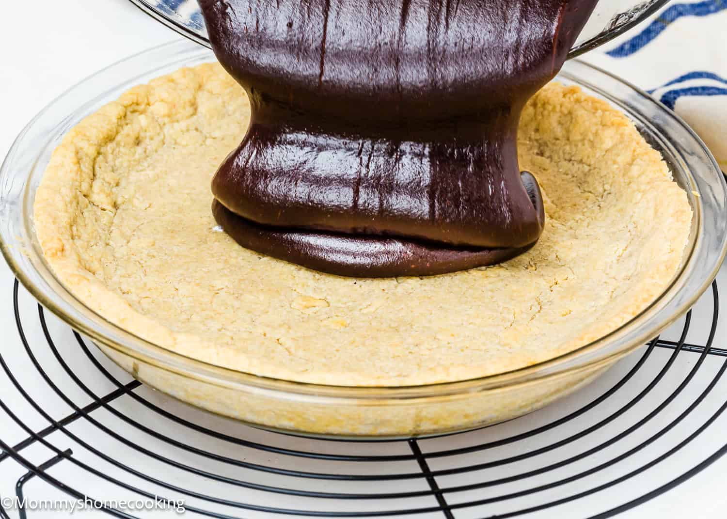 Eggless Easy Brownie Pie filling is being poured into a partially baked pie crust in a glass pie dish on a cooling rack.