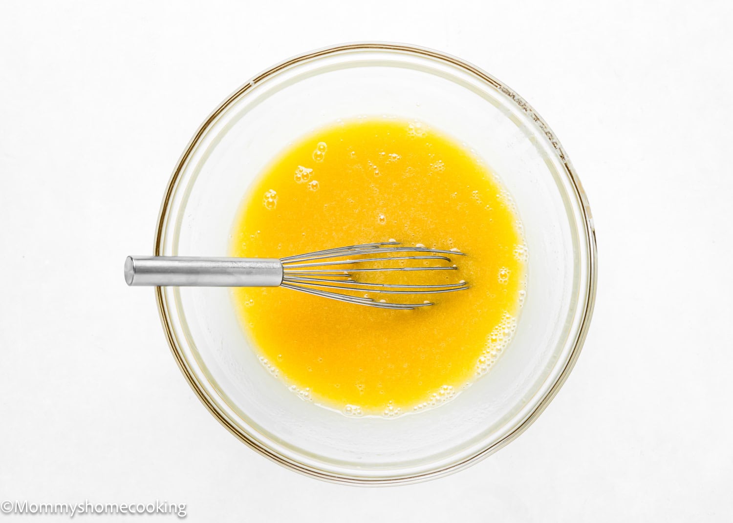 A glass bowl containing melted butter with a metal whisk inside on a pristine white surface.