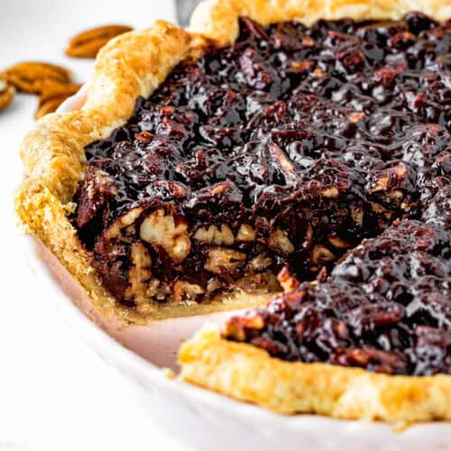 A close-up of an easy chocolate pecan pie with a slice missing, revealing the nutty, chocolate-infused filling. The crust is golden brown, and the pie sits invitingly in a white dish.