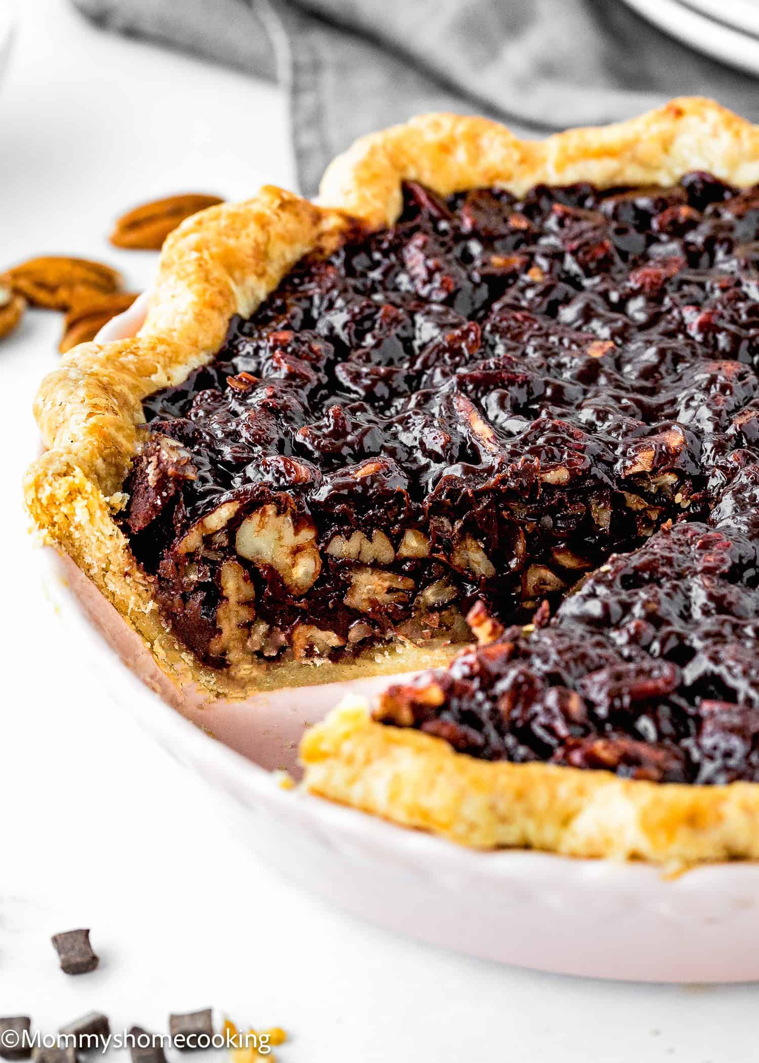 A close-up of an egg-free easy chocolate pecan pie with a slice missing, revealing the nutty, chocolate-infused filling. The crust is golden brown, and the pie sits invitingly in a white dish.