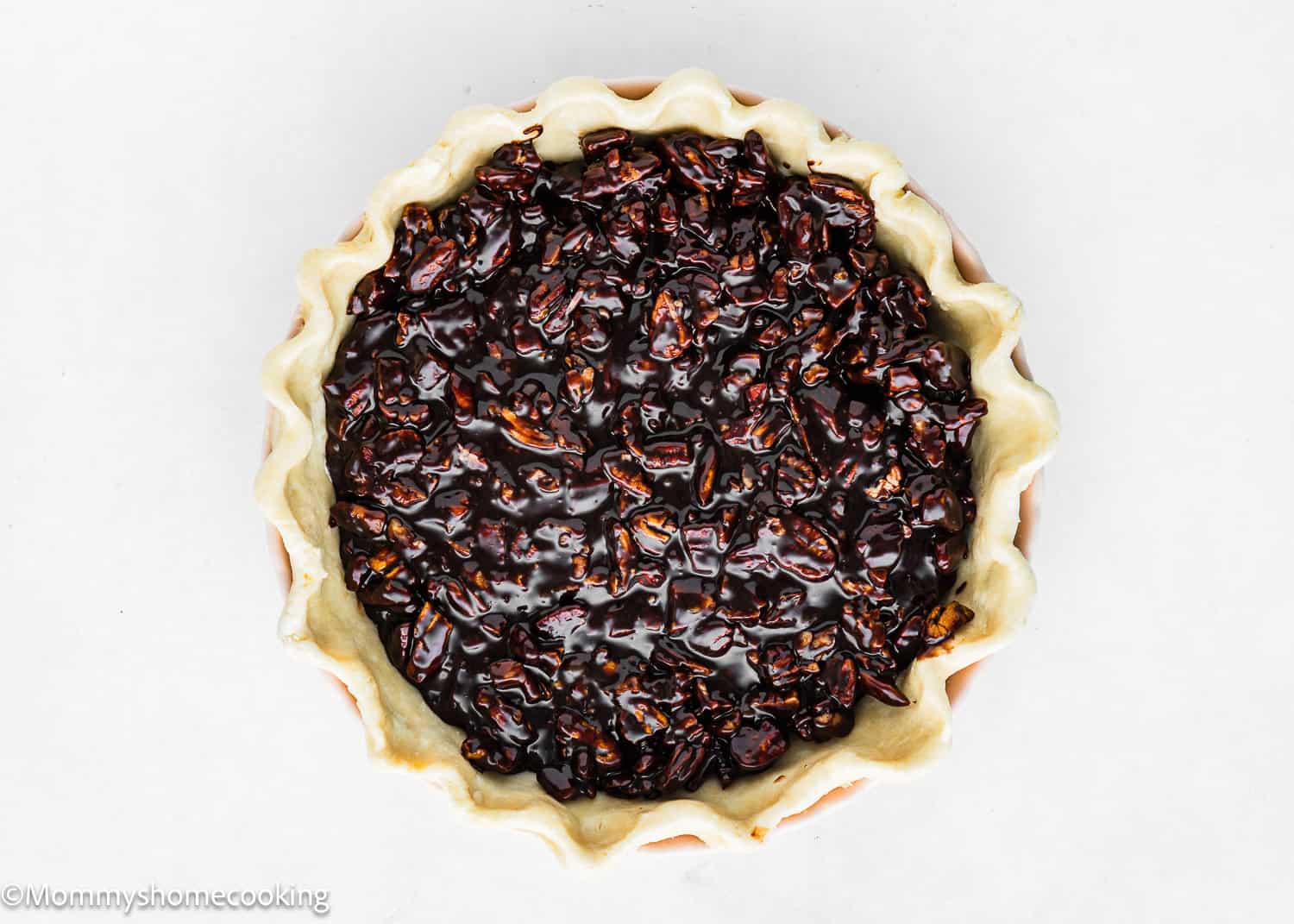 Top view of an unbaked easy, eggless chocolate pecan pie with a crimped edge crust, filled with glossy, dark filling and pecan pieces.