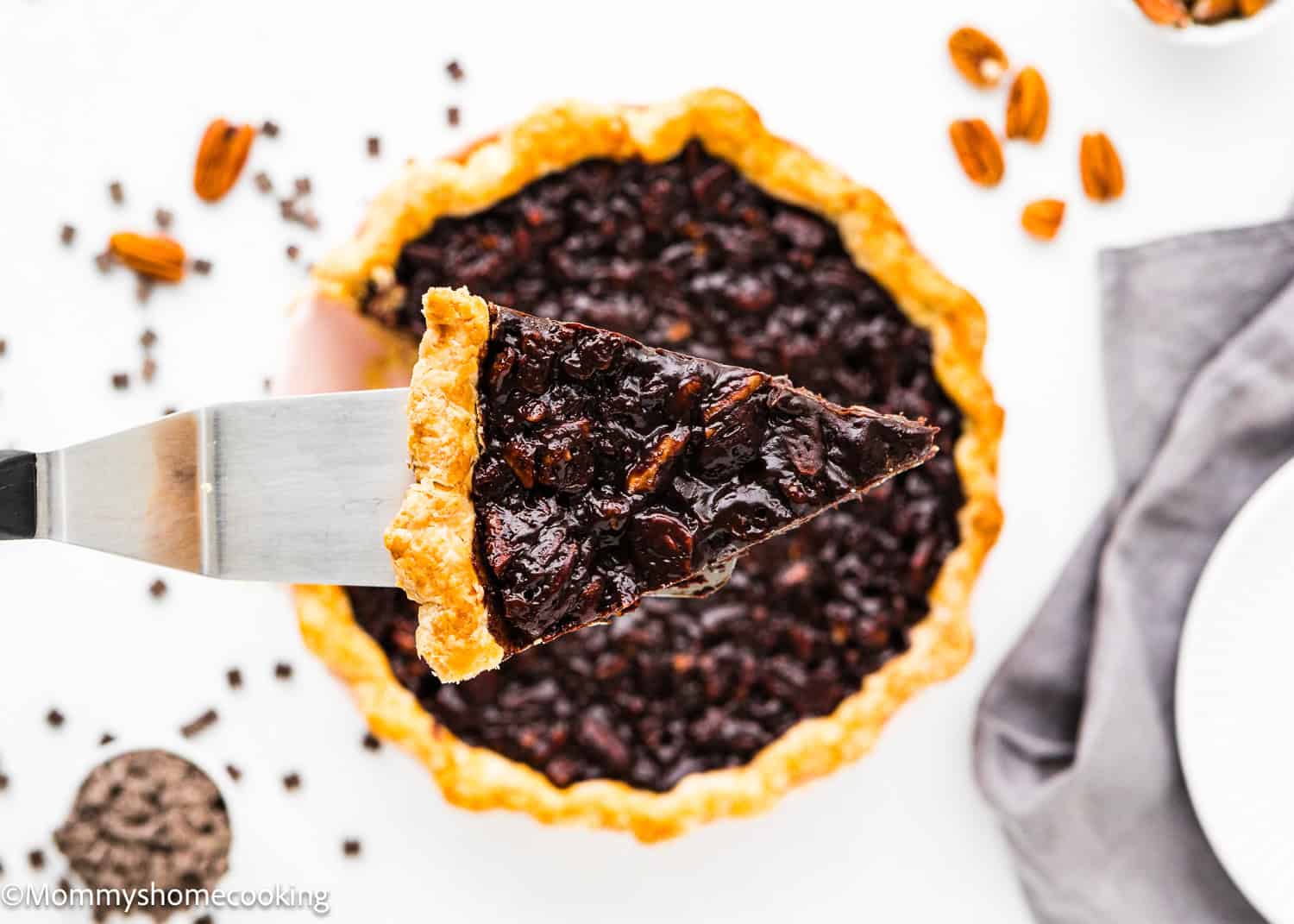 An easy slice of eggless chocolate pecan pie is being lifted from a whole pie on a white surface, with scattered chocolate chips and pecans adding to the allure.