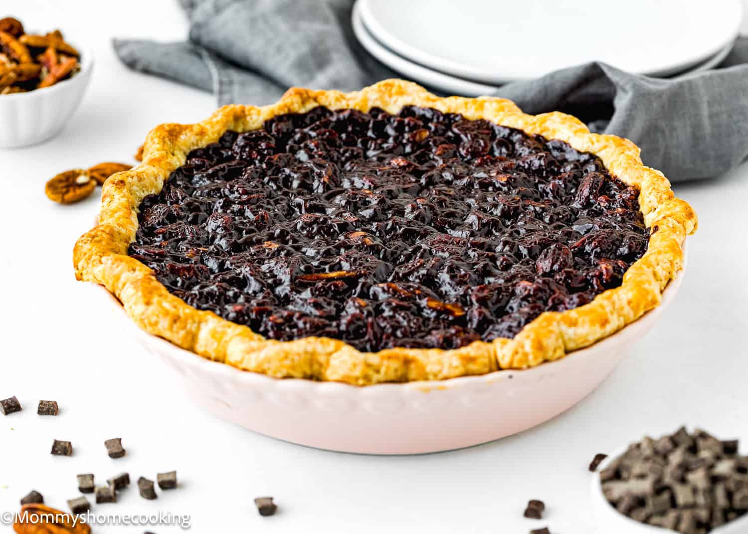 An easy eggless pie with a golden crust, this chocolate pecan delight rests on a table adorned with a grey napkin, plates, and bowls of chopped chocolate and pecans nearby.