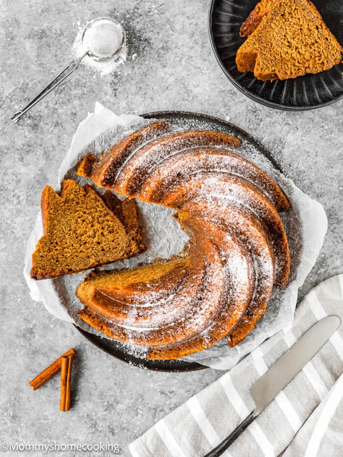 An easy, eggless gingerbread bundt cake sits sliced on a plate, dusted with powdered sugar. Cinnamon sticks and a small sifter with sugar complement the scene, alongside a striped napkin.