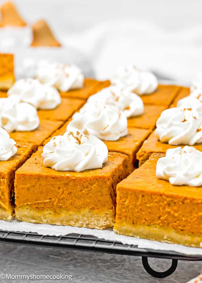 Eggless slices of pumpkin pie with whipped cream topping are elegantly arranged on a cooling rack, making these treats as easy to enjoy as pumpkin pie bars.