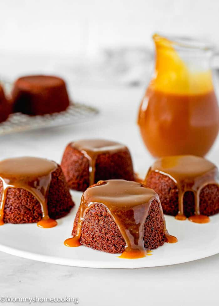 Four Eggless Easy Sticky Toffee Pudding topped with toffee sauce are elegantly arranged on a white plate, with a tempting jar of sticky toffee pudding sauce in the background.